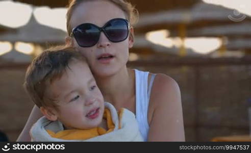 Tilt shot of a lovely family on the beach. Mother and son talking, mom embracing the child wrapped in the towel