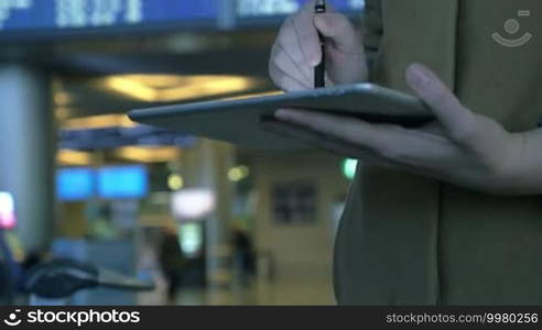 Tilt shot of a woman with tablet computer against flight schedule background. She using pen to type a message