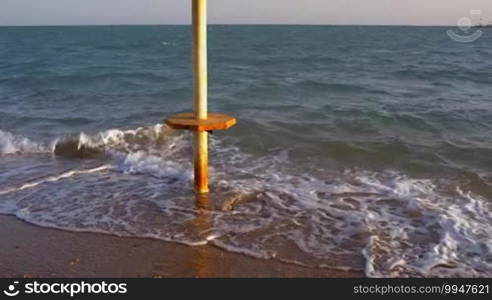 Tilt shot of straw sun umbrella standing lonely on the beach with sea waves washing it