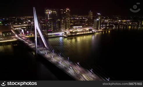 Timelapse shot of transport traffic in Erasmus Bridge and ship sailing along the river on background of illuminated buildings of Rotterdam at night