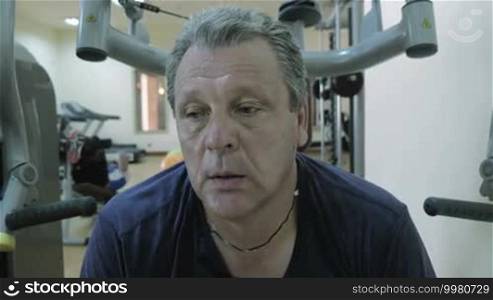 Tired sweaty man having a break and catching breath between sets on exercise machine. Young man doing sit-ups in background