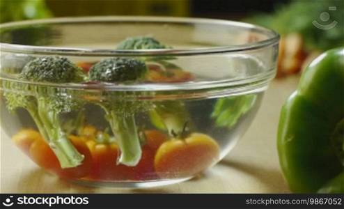Tomatoes and broccoli being washed in a bowl of water, with peppers and other vegetables on the kitchen table. Dolly shot