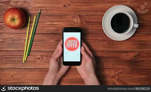 Top view of woman's hands holding smartphone with different sale offers on red tag icon displaying on screen over wooden table background. Discount messages, special limited time offers and last chance to buy on cell phone screen.
