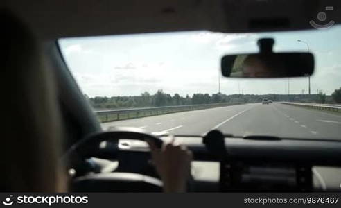 Tourist woman driving car on motorway during summer road trip. View from auto's interior. Female driver controlling vehicle on high speed on curvy asphalt road over beautiful rural scene background.