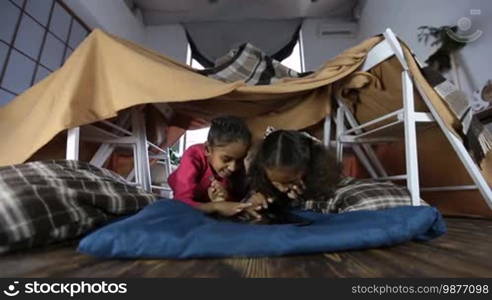 Two attractive mixed race girls having fun and playing online games with a digital tablet as they lie in a cubby house made of chairs and blankets. Excited African American little sisters using a touchpad PC in a domestic room. Low view.