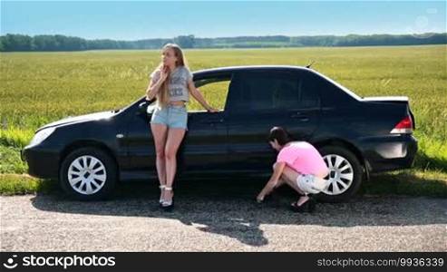 Two attractive women on high heels stopped on a roadside of a rural road during a road trip because the tire is punctured. A cute girl is lifting up the car using a screw jack to change the flat tire with a spare one.