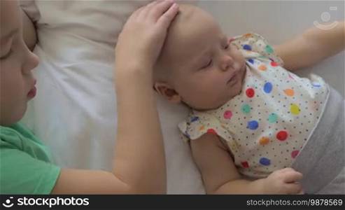 Two children are chilling on a white bed. A seven-year-old boy is taking care of his six-month-old baby sister. She is lying close to him, wearing a colorful polka dot shirt and looking a little bit grumpy. The boy is softly stroking her head and gently blowing on it