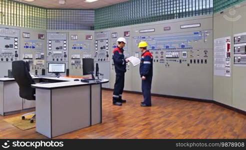 Two engineers talk while looking into logs and giving explanation about circuit at main control panel of gas compressor station