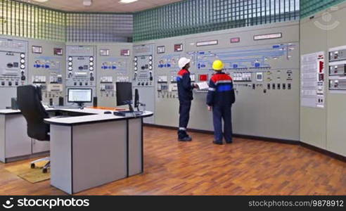 Two engineers talk while showing examples on circuit at main control panel of gas compressor station