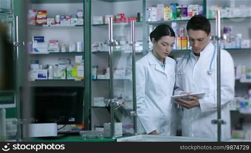 Two experienced pharmacists using a digital tablet while checking medicines in the pharmacy. The male druggist with a stethoscope and his beautiful female colleague are standing in front of shelves with drugs, consulting each other using a touchpad PC in the drugstore.