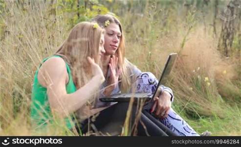 Two female friends sitting on the grass and talking online using a laptop.
