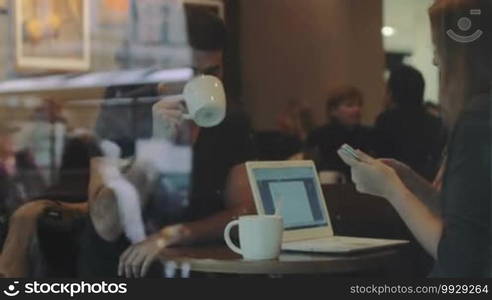 Two friends are sitting in a cafe having a lively conversation. A woman is holding a smartphone, and a laptop is in front of her. The shot is taken through the cafe's window.