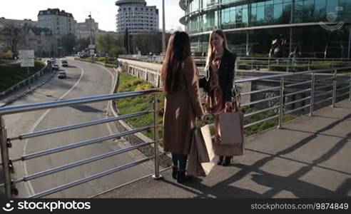 Two multiethnic cute girlfriends with shopping bags chatting while standing on pedestrian bridge at sunset after day shopping over modern cityscape background. Teenage girls discussing their purchases and sharing impressions about new trade centre.