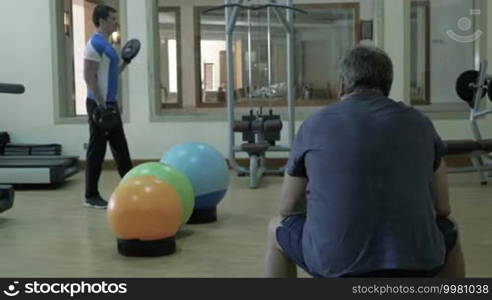 Two people in the gymnasium. Young sportsman exercising with small weight plates while a mature man is taking a break sitting on the bench