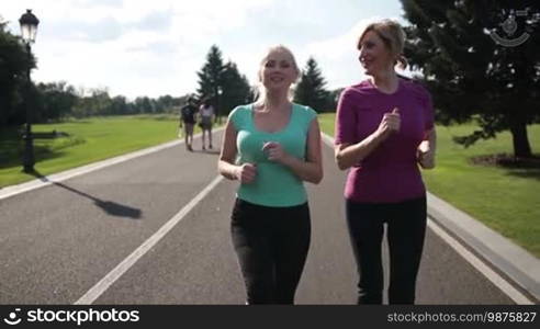 Two smiling athletic senior women running in park on a sunny day. Beautiful adult blonde female runners jogging during outdoor workout in park. Running fit females in sport clothes enjoying training outdoors. Slow motion. Steadicam stabilized shot.