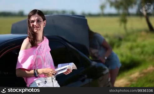 Two young women are trying to apply the screw-jack with help of user guide because their car is broken down on road.