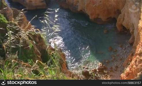 View from a high, steep cliff on a rocky bay surrounded by bare cliffs; the sun glitters in the shallow turquoise water; Coast of Algarve, Portugal.
