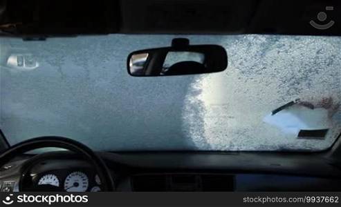 View from inside of car's cab. Female driver scraping the car windscreen with ice scraper. Woman cleaning car from snow with brush on winter morning.