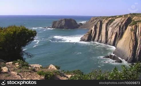 View of high cliffs / the sea from a viewpoint with green bushes; rocks in the sea, coast of the Algarve in Portugal; blue sky.