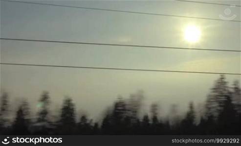 View of passing landscape while traveling by train with trees and bright evening sun shining between electric wires
