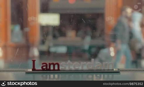 View of small plastic figure of Iamsterdam letters sculpture on the scratched windows on tram against blurred cityscape with buildings and road and bikes, Amsterdam, Netherlands