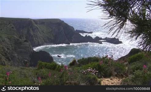 View of the ocean / high cliffs / a bay from a colorful flower meadow; rocks in the sea, coast of the Algarve in Portugal; the sun glistens in the water, blue sky.