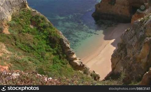 Weißer verlassener Natursandstrand vor türkisblauem Wasser zwischen hohen, steilen, teilweise grün bewachsenen sandsteinfarbenen Felsen / Klippen - Küste der Algarve, Portugal.