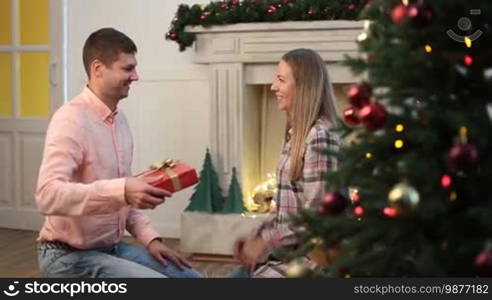 What a great surprise! Beautiful woman with long hair receiving Christmas gift box from beloved husband over xmas decorated room. Affectionate man surprising his young wife with present during winter holidays. Dolly shot.