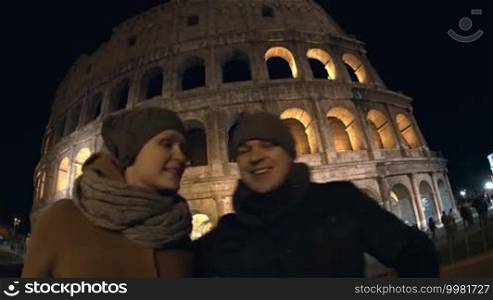 Wide angle shot of young loving couple making selfie with mobile phone by the Coliseum at night. Happy tourists kissing and leaving