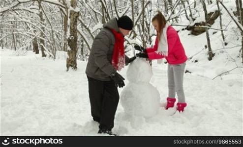 Winter holidays in a snowy forest, teenage couple making a snowman