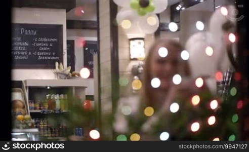 Woman drinking tea in a cafe and looking at Christmas lights. View through twinkling garland. Defocus at the beginning.