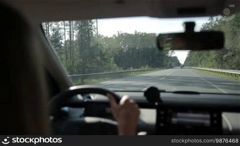 Woman driving car on motorway through the forest in sunlight. View from vehicle's interior. Car traveling on asphalt highway over beautiful landscape background.