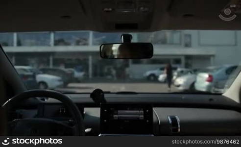 Woman driving car to exit parking lot near shopping mall with blurred trade center in the background. View from vehicle's interior. Car leaving open parking area next to trade center.