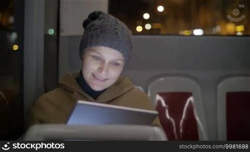 Woman in coat and stocking cap chatting on tablet computer during city bus ride