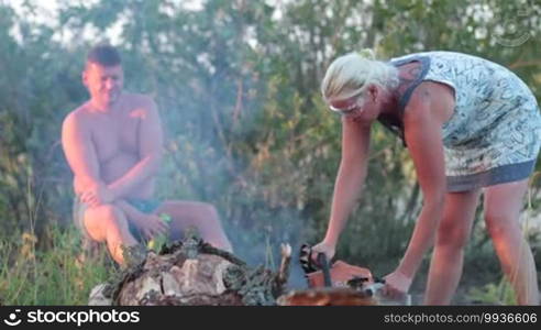 Woman in glasses sawing thick dry tree with chainsaw. Man with can of beer sitting on stump and watching. Happy family picnicking on summer vacation