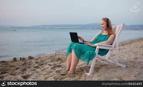 Woman relaxing at the beach in a deckchair alongside the ocean with her laptop computer and wearing a headset talking with her friend with Skype