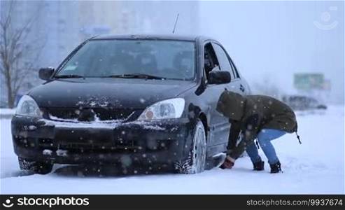 Woman shoveling and removing snow from her car's wheel. The car stuck in the snow. Young lady in warm clothes digging car out of the snow during snowfall.