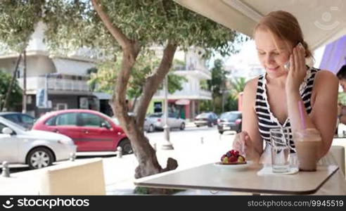 Woman sitting at an urban cafeteria alongside a street talking on a mobile phone with a serious expression as she listens to the conversation