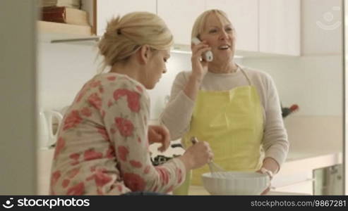 Women in kitchen at home, mother speaking on the phone while daughter helps her cooking
