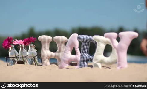 Word "happy" made of fabric padded letters and flowers in glass bottles on the sand at the beach over blurred background of kid running on shore.