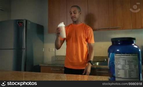 Young African American man in domestic kitchen, drinking a milkshake with proteins and vitamins. The guy tastes the beverage and smiles