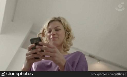 Young beautiful Caucasian blonde woman typing email message on smartphone and laughing. Low angle view, dolly shot