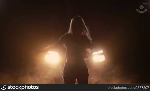 Young beautiful excited woman listening to music and dancing with long blond hair flying in the bright car headlights at night. Overjoyed female in shorts enjoying leisure dancing in clouds of smoke over yellow car headlights at night. Slow motion