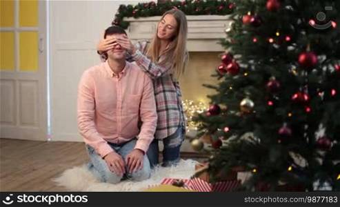 Young beautiful wife keeping her husband's eyes covered with hands while cute daughter giving Christmas gift in Christmas decorated room. Adorable little girl giving present to her handsome father during family winter holidays at home. Dolly shot.