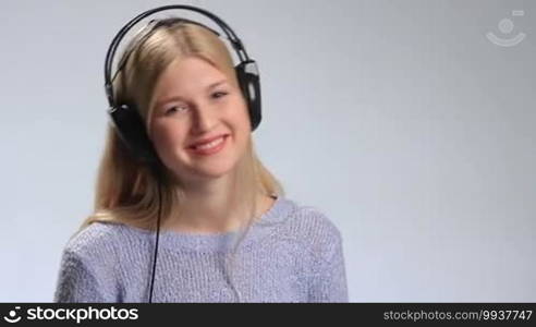 Young beautiful woman enjoying music with headphones on white. Cheerful teenage girl in black headphones touching the earcups with hand, swaying her head to the rhythm of song and smiling into the camera