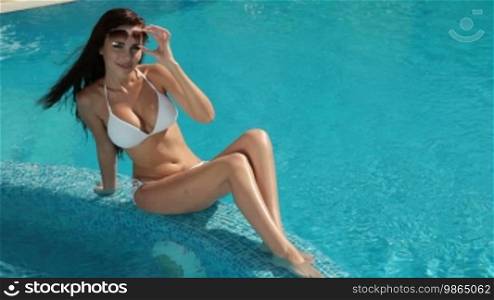 Young bikini woman sunbathing by swimming pool, looking at camera