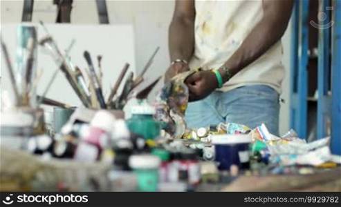 Young black people painting for hobby. African American man working as a professional painter, skillful Cuban artist in an art studio, cleaning brush and tools on a table full of equipment, canvas, and paint