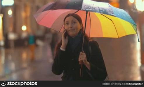 Young brunette woman stands with multicolored umbrella in her hand on the street on a rainy day and talks on the phone