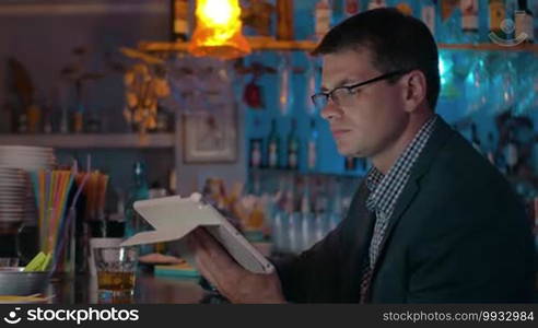 Young businessman in glasses and suit using tablet computer while sitting at the bar counter and having alcoholic drink