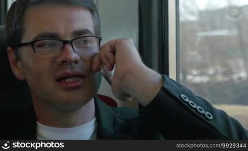 Young businessman is sitting in a train and talking on the phone. After the end of the conversation he's looking out the window.